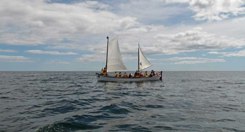 From a distance, a sailboat with white sails floats on water under a gray-blue sky.